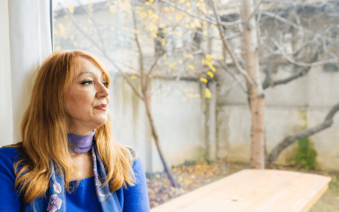 Middle aged woman with red hair looking out the window. She is wearing a blue sweater and floral scarf.